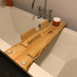 May include: A light brown wooden bathtub tray with a natural live edge, featuring a vertical book or tablet stand, a small slot for a stemware glass, and a circular cutout. The tray rests across a white bathtub with a chrome faucet fixture.