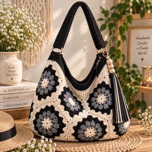 May include: A crochet hobo bag featuring a black, gray, and cream granny square pattern. The bag has a thick black crocheted handle, gold-tone metal rings, and a decorative tassel with three wooden beads attached to the side of the bag.