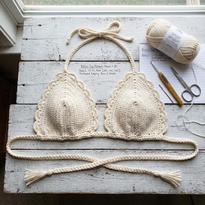 May include: A cream-colored crochet bikini top with scalloped edges and braided ties featuring tassels. Beside it lies a ball of sport weight cotton yarn, a crochet hook, scissors, a needle, and a paper with the text Bikini Top Pattern.