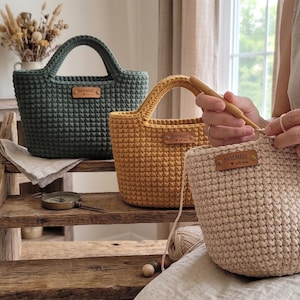 Op de afbeelding: Drie gehaakte tassen in donkergroen, mosterdgeel en beige, elk voorzien van een klein rechthoekig leren label met de tekst HANDMADE en een hartje. De beige tas wordt bewerkt met een houten haaknaald en bijpassend garen.