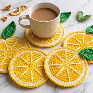 Puede incluir: Un juego de cuatro posavasos redondos de ganchillo con forma de rodaja de limón en amarillo y blanco, cada uno con una hoja verde tejida en el borde superior. Una taza de cerámica beige con una bebida marrón claro descansa sobre uno.