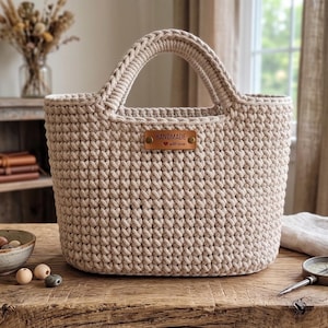 Op de afbeelding: Een beige gehaakte tas met twee ge&iuml;ntegreerde handvatten en een rechthoekig bruin leren label aan de voorzijde met de tekst "HANDMADE with love". De tas heeft een gestructureerd, herhalend steekpatroon en een brede opening.