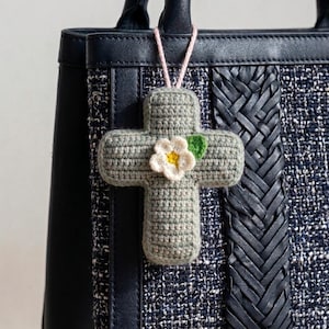 May include: A sage green crocheted cross ornament featuring a small white flower with a yellow center and a single green leaf. The cross is attached to a light pink string and hangs from the handle of a dark navy blue textured handbag.