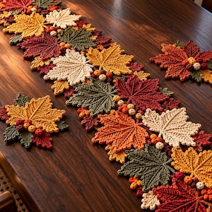 Könnte beinhalten: Ein gehäkelter Tischläufer und zwei passende Untersetzer mit einem Muster aus Ahornblättern in Creme, Gelb, Orange, Rot und Olivgrün, akzentuiert durch kleine gehäkelte Beerengruppen, die zwischen den strukturierten Blattformen liegen.
