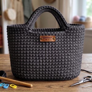 Puede incluir: Un bolso de ganchillo gris oscuro con dos asas integradas y una etiqueta rectangular de cuero marrón centrada en la parte delantera que dice HANDMADE with love. Un ganchillo de madera, tres marcadores de puntos y tijeras están cerca.