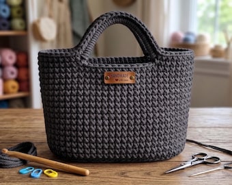 May include: A dark grey crocheted tote bag with two integrated handles and a rectangular brown leather tag centred on the front that reads HANDMADE with love. A wooden crochet hook, three plastic stitch markers, and metal scissors are nearby.