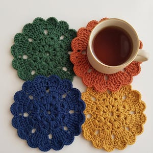 May include: Four handmade crochet coasters in shades of green, orange, navy blue, and gold. A beige teacup filled with tea sits on the orange coaster. The coasters have a scalloped edge and a floral design.