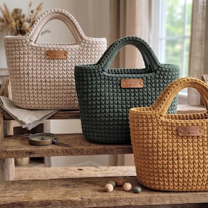 Op de afbeelding: Drie gehaakte handtassen in beige, donkergroen en mosterdgeel. Elke tas heeft een gestructureerde vorm, twee stevige handvatten en een klein rechthoekig label in lederlook aan de voorzijde met de tekst Handmade with love erop.
