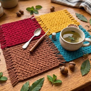 May include: Four square crochet coasters in red, yellow, brown, and teal, each featuring a textured stitch pattern and fringed edges. A silver spoon, a wooden crochet hook, a metal thimble, and a ceramic mug of tea are placed on the coasters.