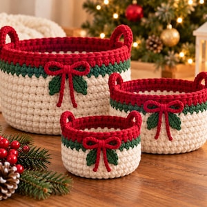 Puede incluir: Un conjunto de tres cestas redondas de ganchillo de diferentes tamaños, con una base de color crema, una banda central verde oscuro y un borde superior rojo. Cada cesta incluye dos asas laterales y un lazo decorativo de hilo rojo.
