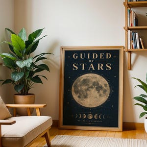 Könnte beinhalten: Ein gerahmtes Poster mit Holzrahmen zeigt ein himmlisches Design. Das Poster zeigt eine detaillierte Illustration des Mondes, umgeben von Sternen, und die Worte "GUIDED BY STARS". Das Poster wird in einem Raum mit Pflanzen und einem Holzstuhl ausgestellt.