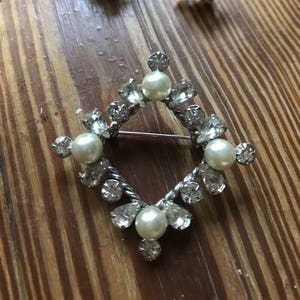 May include: A silver-toned jewelry set consisting of a diamond-shaped brooch and a pair of matching screw-back earrings. Each piece features a central faux pearl surrounded by clear, faceted rhinestones in round and pear-shaped settings.