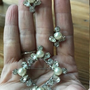 May include: A jewelry set consisting of a diamond-shaped brooch and two matching clip-on earrings. Each piece features a silver-toned metal frame set with clear faceted rhinestones and a central round white faux pearl accent.