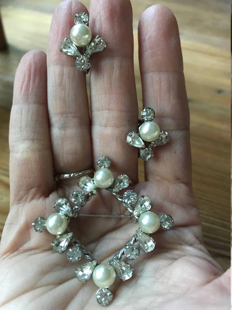 May include: A jewelry set consisting of a diamond-shaped brooch and two matching clip-on earrings. Each piece features a silver-toned metal frame set with clear faceted rhinestones and a central round white faux pearl accent.