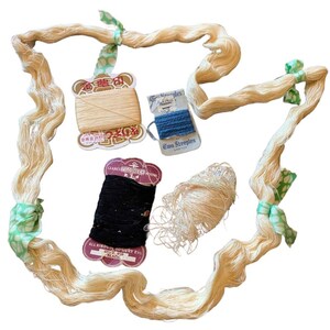 May include: A collection of sewing supplies including a long, continuous strand of cream-colored thread tied with green patterned fabric strips, a cream thread spool, a blue thread spool labeled Two Steeples, a black thread spool, and a loose ball of cream thread.