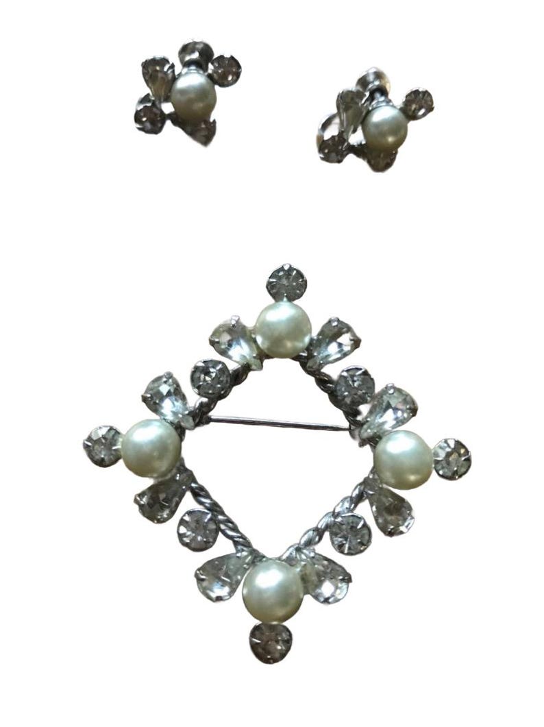 May include: A jewelry set consisting of a diamond-shaped brooch and a pair of matching stud earrings. Each piece features a central cream-colored faux pearl surrounded by clear, faceted crystals set in a silver-toned metal frame.