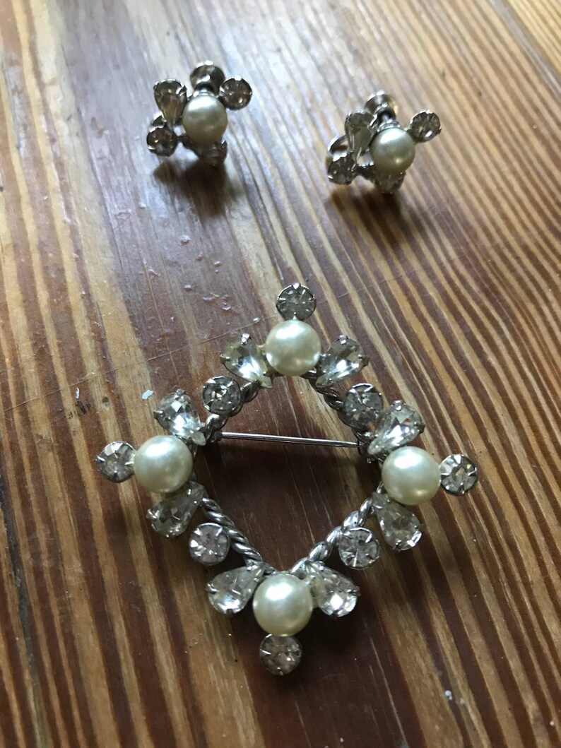 May include: A silver-toned jewelry set consisting of a diamond-shaped brooch and a pair of matching screw-back earrings. Each piece features a central faux pearl surrounded by clear, faceted rhinestones in round and pear-shaped settings.