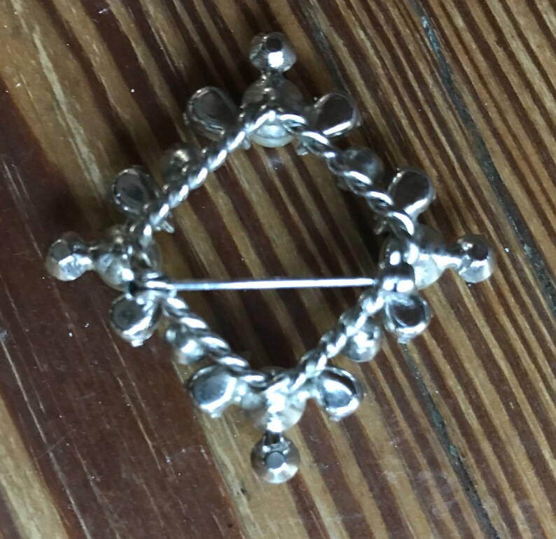 May include: A silver-toned metal brooch in a diamond shape, featuring a twisted wire frame adorned with small, round, clear faceted stones set at regular intervals along the perimeter and at each of the four points of the diamond structure.