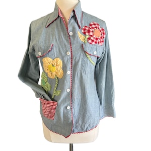May include: Light blue long-sleeve shirt with floral applique details. The shirt features red gingham trim along the collar, cuffs, and pockets. Two flower appliques are on the front, one red and one yellow. The shirt has white buttons.