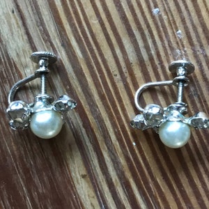 May include: A pair of silver-toned screw-back earrings featuring a central round white faux pearl surrounded by small clear faceted stones. The metal hardware includes a threaded screw mechanism for adjustment, providing a secure fit for the earlobe.