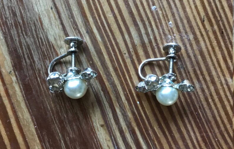 May include: A pair of silver-toned screw-back earrings featuring a central round white faux pearl surrounded by small clear faceted stones. The metal hardware includes a threaded screw mechanism for adjustment, providing a secure fit for the earlobe.
