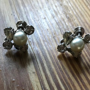 May include: A pair of silver-toned screw-back earrings featuring a central white faux pearl surrounded by three small clear faceted stones arranged in a cluster. The metal setting has a polished finish and the screw mechanism is visible at the back.