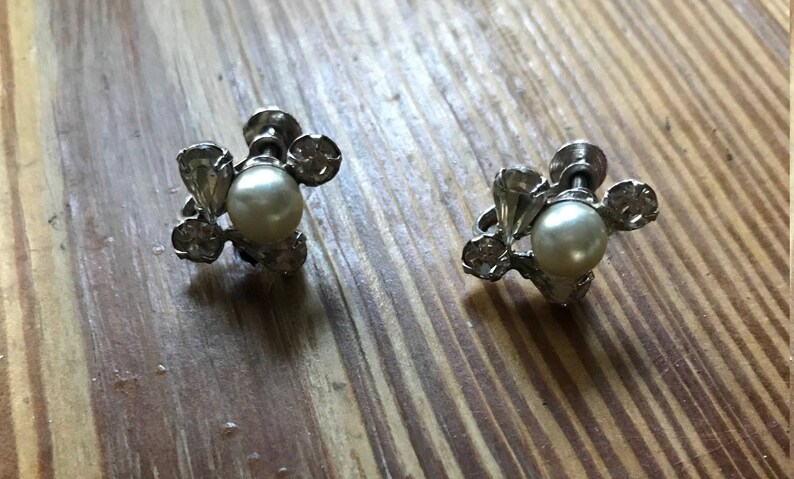 May include: A pair of silver-toned screw-back earrings featuring a central white faux pearl surrounded by three small clear faceted stones arranged in a cluster. The metal setting has a polished finish and the screw mechanism is visible at the back.
