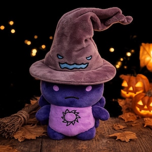 May include: A plush toy featuring a dark purple body with a light purple belly displaying a black sun emblem. The toy wears a large, soft, dark purple witch hat with an embroidered, light blue, frowning face on the front brim and two small eyes.