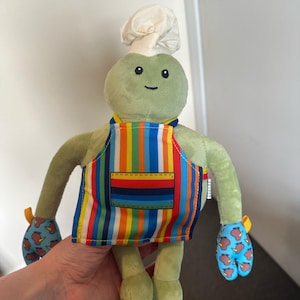 May include: A plush toy shaped like a green creature wearing a white chef hat, a colorful vertical striped apron with a central pocket, and blue oven mitts featuring a repeating pattern of small brown kiwi birds on each hand.