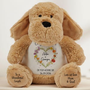 May include: A tan plush dog toy with a white chest panel featuring a floral heart design, the names Anton & Georgia, and the text ON YOUR WEDDING DAY 26.09.2026. The feet are embroidered with To a Wonderful Couple and Lots of love Mum & Dad.