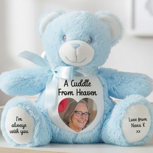 May include: A light blue plush teddy bear featuring a white chest patch with the text A Cuddle From Heaven above a heart-shaped photograph of a smiling person with glasses. The bear has a light blue ribbon and text on its feet reading I'm always with you and Love from Nana K xx.