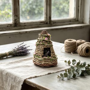 May include: A conical beehive skep crafted from woven natural fibers, featuring a small rectangular entrance. The structure is decorated with thin green vines and small pink fabric flowers wrapped around the exterior in a spiral pattern.