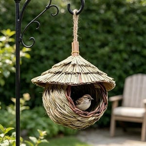 May include: A woven straw birdhouse with a conical thatched roof and a circular opening, suspended by a jute rope from a black metal shepherd's hook. A small brown and white sparrow is perched inside the nest, which features a rounded base.