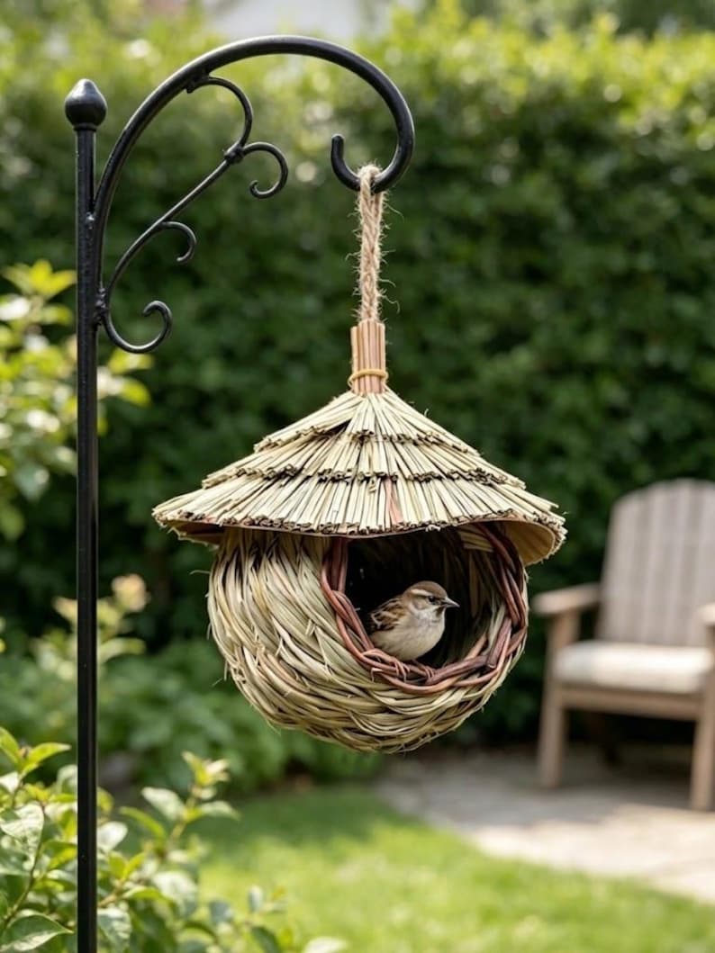 May include: A woven straw birdhouse with a conical thatched roof and a circular opening, suspended by a jute rope from a black metal shepherd's hook. A small brown and white sparrow is perched inside the nest, which features a rounded base.