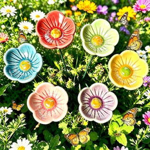 May include: A set of six ceramic flower-shaped garden stakes in red, light green, blue, yellow, pink, and purple, each featuring a raised center and thin metal stem, arranged among various colorful garden flowers and monarch butterflies.