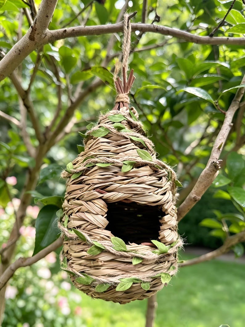 May include: A teardrop-shaped birdhouse woven from light tan dried grass and straw. The structure features a circular entrance hole and is wrapped with a thin jute twine intertwined with small, artificial green fabric leaves for decoration.