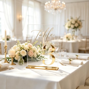 May include: A clear acrylic table number sign featuring the gold script text Table 3. The rectangular sign is supported by a gold metal base and is positioned on a white tablecloth, serving as a centerpiece for a formal event setting.