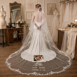 May include: A bride in a white satin wedding dress stands with her back to the camera, wearing a long veil with floral lace trim. The veil trails onto the floor, with a photo of the couple and the date "October 8, 2026" at the bottom.