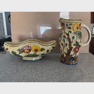 May include: A decorative ceramic pitcher and bowl set. The pitcher has a handle and is adorned with a floral and tree branch design in shades of yellow, red, and blue. The bowl has a wavy rim and a similar floral pattern.