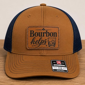 May include: A tan and navy blue trucker hat with a rectangular patch on the front. The patch features the text "Bourbon helps" in black lettering alongside an illustration of a glass with ice cubes. A black sticker is on the brim.