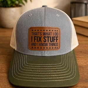May include: A gray and olive green trucker hat with a mesh back. The hat features a brown leather patch with the text "THAT'S WHAT I DO I FIX STUFF AND I KNOW THINGS" in white lettering. The hat has a curved brim.