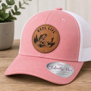 May include: A pink and white trucker hat with a leather patch that says "REEL LIFE" with a graphic of a fish jumping out of the water. The hat has a curved brim and a mesh back. The hat is small/medium.