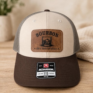 May include: A beige and dark brown trucker hat with a mesh back. A rectangular brown leather patch on the front features the text BOURBON, an illustration of a glass with ice, and the text Life is Better with Bourbon. A black sticker reads RICHARDSON EST. 1970 LOW-PRO 115 TRUCKER MD/LG.