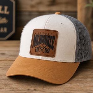May include: Baseball cap with a brown brim, white front panel, gray mesh back, and a brown leather patch. The patch features the text "Baseball Milwaukee 1969" with crossed bats and a baseball.