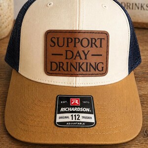 May include: A trucker hat with a cream-colored front panel, a brown brim, and dark blue mesh back panels. A rectangular brown leather patch on the front features the black text SUPPORT - DAY - DRINKING. A black sticker on the brim reads RICHARDSON EST. 1970 ORIGINAL 112 TRUCKER ADJUSTABLE.