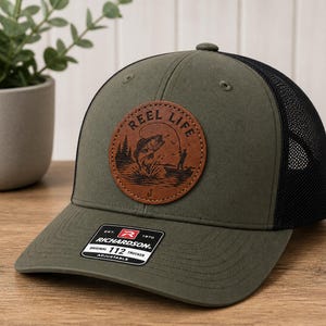 May include: Olive green trucker hat with a black mesh back. The front features a brown leather patch with the words "REEL LIFE" and a fishing scene. The hat has a curved brim and an adjustable strap.