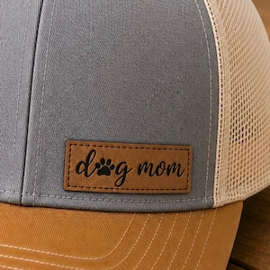 May include: A gray and tan trucker hat with a brown brim. The hat features a patch with the words "dog mom" and a paw print. The hat has a mesh back and a button on top.
