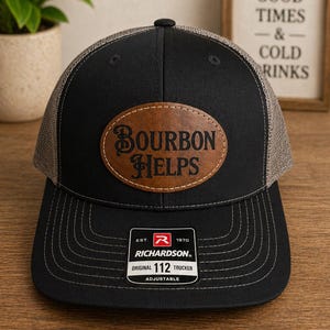 May include: Black trucker hat with a brown leather patch that reads "BOURBON HELPS." The hat has a mesh back and a curved brim. The Richardson brand label is visible. A sign in the background says "GOOD TIMES & COLD DRINKS."
