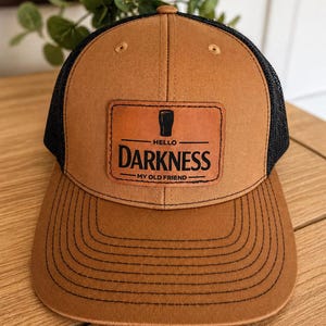 May include: A brown and black trucker hat with a curved brim. The hat features a leather patch with the words "HELLO DARKNESS MY OLD FRIEND" and a beer glass graphic. The hat has a mesh back.