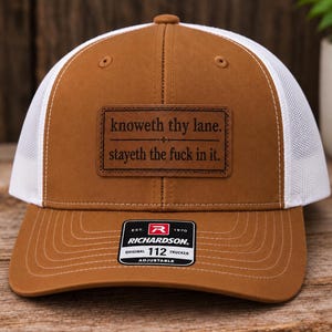 May include: A brown and white trucker hat featuring a rectangular leather patch on the front. The patch is engraved with the text "knoweth thy lane. stayeth the fuck in it." A black and white sticker on the brim displays the Richardson 112 logo.
